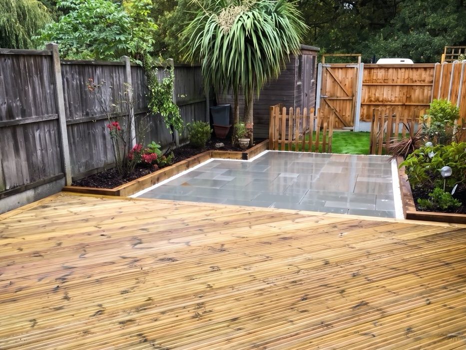 New timber decking leading to a grey slab patio, with planted borders along the fence