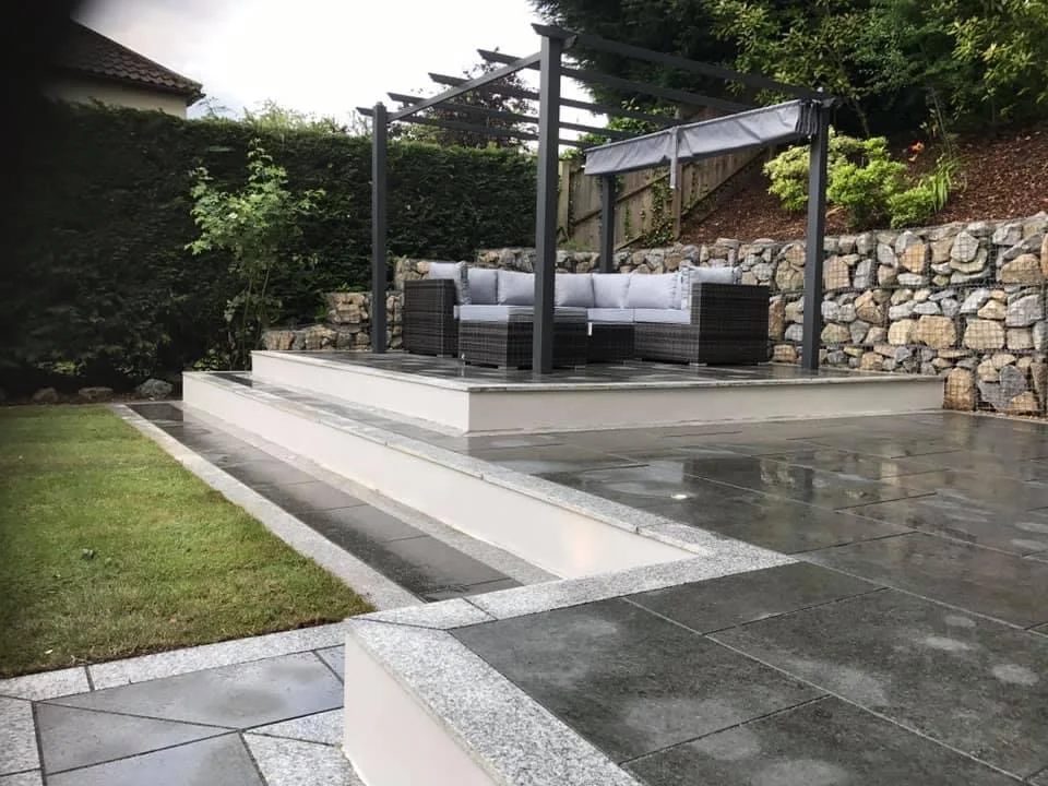 Raised dark stone patio with granite edging, a pergola and a gabion wall